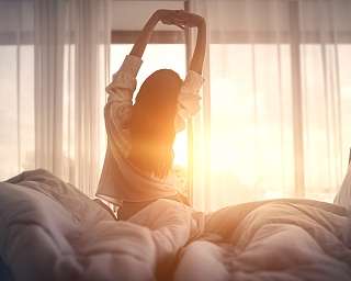 Eine Frau streckt sich achtsam im Bett beim Sonnenaufgang – Morgenritual voller Ruhe und Licht in einem Wohlfühlhotel bei WUNDERORT