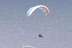 Tandem-Paraglider schwebt über dem Gardasee mit Blick auf Ausflugsschiff und bewaldete Ufer – ideal für Sport- und Abenteuerurlaub in Italien I WUNDERORTE