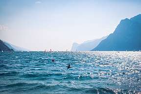 Blick über den Gardasee mit zahlreichen Windsurfern und Segelbooten vor sonniger Bergkulisse – beliebtes Revier für Wassersport und Natururlaub I WUNDERORTE
