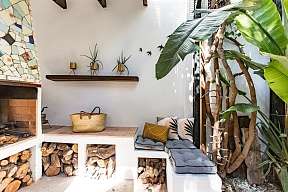 Outdoor Lounge mit Kamin und Bananenpflanzen, gestapeltes Holz unter der Sitzbank mit gemütlichen hellblauen Kissen - entspannter Rückzugsort