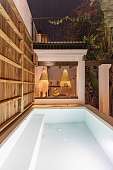 Beleuchteter Pool bei Nacht mit überdachtem Lounge-Bereich &amp; dekorativen Lampen aus Bast