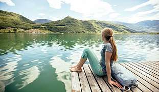Frau genießt sitzend auf einem Steg eine Wander-Pause und schaut zufrieden oder verträumt über einen See.