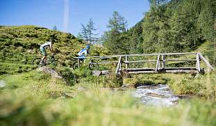 Zwei Radfahrer überqueren eine Holzbrücke inmitten einer Berglandschaft bei Sonnenschein