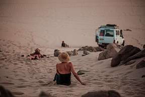 Frau sitzt im Sand einer ägyptischen Wüste mit Blick auf einen Geländewagen – achtsamer Wüstenausflug mit dem Jeep