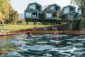 Ein Paar genießt entspannte Momente im Naturpool mit Blick auf moderne Design-Chalets in Südtirol – Erholung in der Natur I WUNDERORTE