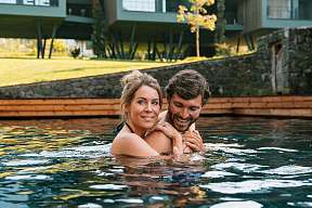 Paar im Naturpool vor modernen Chalet-Suiten – Wellness und Lebensfreude in Südtirol – WUNDERORTE