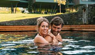 Ein Paar genießt entspannte Zweisamkeit im Naturpool vor modernen Design-Chalets in Tirol – Wellnessurlaub mit Stil I WUNDERORTE