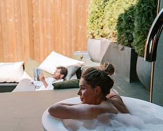Frau entspannt im Outdoor-Whirlpool, im Hintergrund ein Mann beim Lesen auf der Terrasse einer privaten Wellnesssuite – Rückzugsort für Ruhe, Genuss und mentale Erholung mit WUNDERORTE in Südtirol.