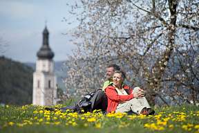 Ein Paar sitzt entspannt auf einer Frühlingswiese mit Löwenzahnblüten und Blick auf die Kirche von Kastelruth – Auszeit in der Natur I WUNDERORTE