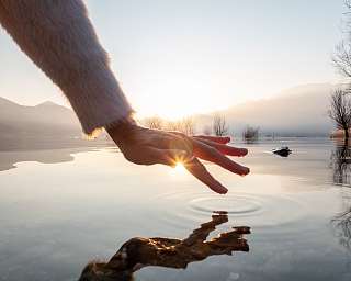 ine Hand berührt im Sonnenaufgang das stille Wasser – ein stiller Moment voller Licht, Ruhe und Achtsamkeit, wie geschaffen für WUNDERORTE.Hand über stillem Wasser – Lichtmoment in der Natur bei WUNDERORTE