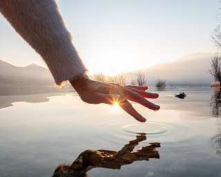 Eine Hand berührt im Sonnenaufgang das stille Wasser – ein stiller Moment voller Licht, Ruhe und Achtsamkeit, wie geschaffen für WUNDERORTE.