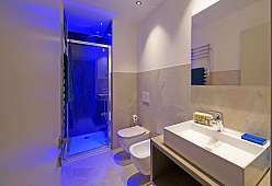 Modernes Bad eines Ferienhauses am Gardasee mit verglaster Dusche in blauem Licht, WC, Bidet sowie großem weißen Waschtisch vor Natursteinfliesen