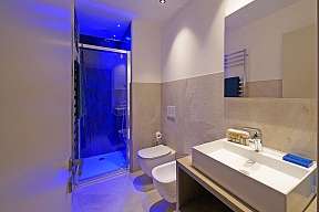 Modernes Bad eines Ferienhauses am Gardasee mit verglaster Dusche in blauem Licht, WC, Bidet sowie großem weißen Waschtisch vor Natursteinfliesen