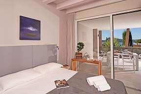 Hell gestaltetes Schlafzimmer der Ferienwohnung Lamasu am Gardasee mit Boxspringbett, Panoramafenster, Seeblick und Zugang zur möblierten Terrasse I WUNDERORTE
