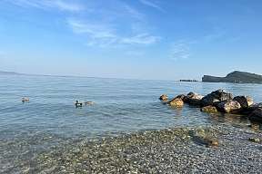 Zwei Enten schwimmen im klaren Wasser vor einem Kiesstrand am Gardasee; rechts liegen Felsbrocken, im Hintergrund erhebt sich die Rocca di Manerba unter blauem Himmel.