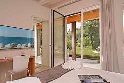 Modernes Schlafzimmer in einem Ferienhaus am Gardasee mit Bett, weißen Handtüchern, TV und offener Terrassentür; dahinter grüner Garten.“