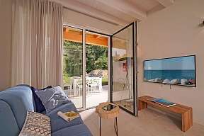Helles Wohnzimmer der Ferienwohnung Lamasu am Gardasee mit Sofa, TV, Zugang zur Terrasse und Blick ins Grüne I WUNDERORTE