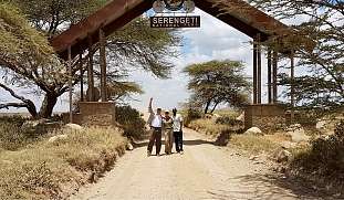 Drei Menschen stehen am Eingangstor des Serengeti Nationalparks unter schattigen Akazienbäumen